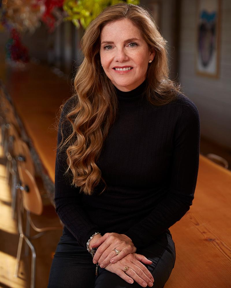 Leslie Chihuly with long, wavy strawberry-blonde hair, wearing a black turtleneck and sitting at the edge of a polished wooden table with her hands clasped in her lap. The background features a long row of chairs and colorful, blurred glass art pieces in a warm, dimly lit room.