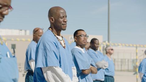 A group of people wearing blue shirts stand in a prison yard, meditating with eyes closed