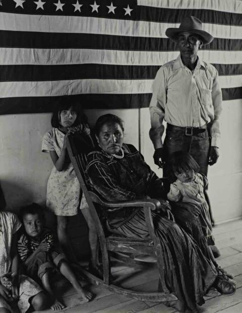 Francis Nakai and Family, 1950, photo by Laura Giplin. Black and white photo of a Navajo family, with a woman sitting in a rocking chair in front of a partial American Flag.