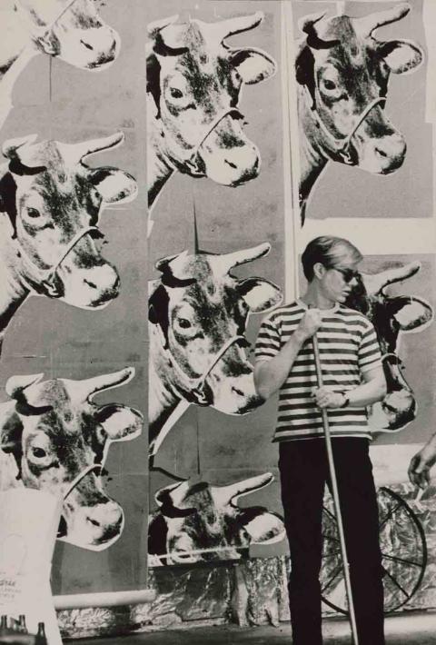 Andy in the Factory with Cow Wallpaper, 1965, photo by Billy Name. A black and white image showing a person wearing a black and white striped shirt, black pants, sunglasses, and a watch holding a long pole with a cow patterned wall in the background