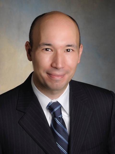 A person with a shaved head, a dark striped suit jacket, and blue striped tie smiles at the viewer.