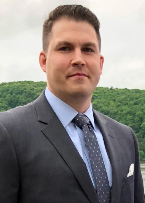 A person with short brown hair and a dark suit and tie looks at the viewer. The person is standing in front of a distant hill covered with trees.