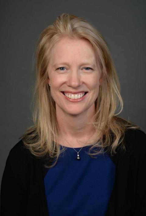 A person with long blond hair, a black jacket, and a blue shirt smiles at the viewer.