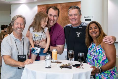 A group of smiling people standing near a small, hightop cocktail table with two glasses of wine on it. One person is holding a child.