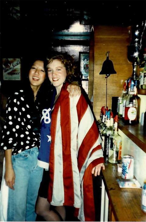 Two students stand next to a shelf with alcohol bottles, arms around each other's shoulders. The photo is somewhat dark and illuminated by flash.