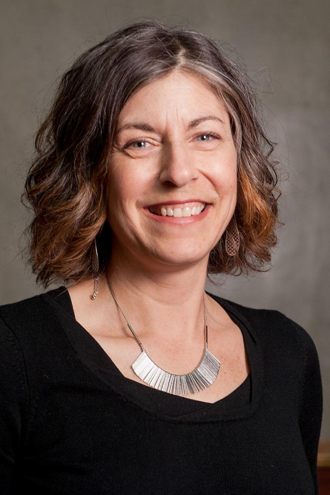 Erin McCloskey smiling with wavy, multi-toned hair, wearing a black top and a striking silver necklace.