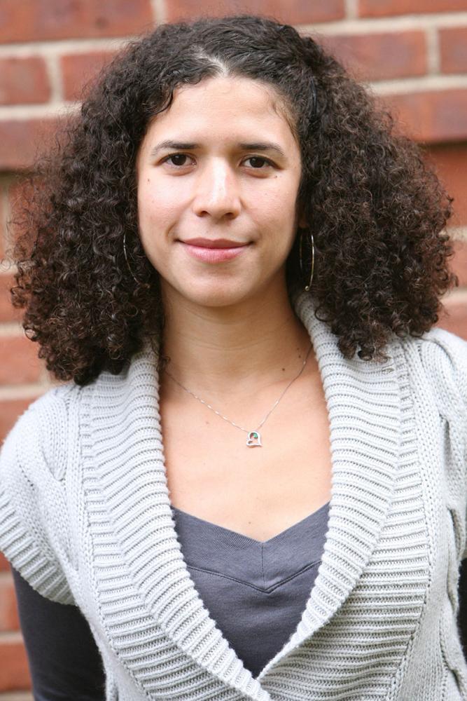 Eve Dunbar smiling with voluminous curly dark hair, wearing a gray knit sweater and a necklace, standing in front of a brick wall.