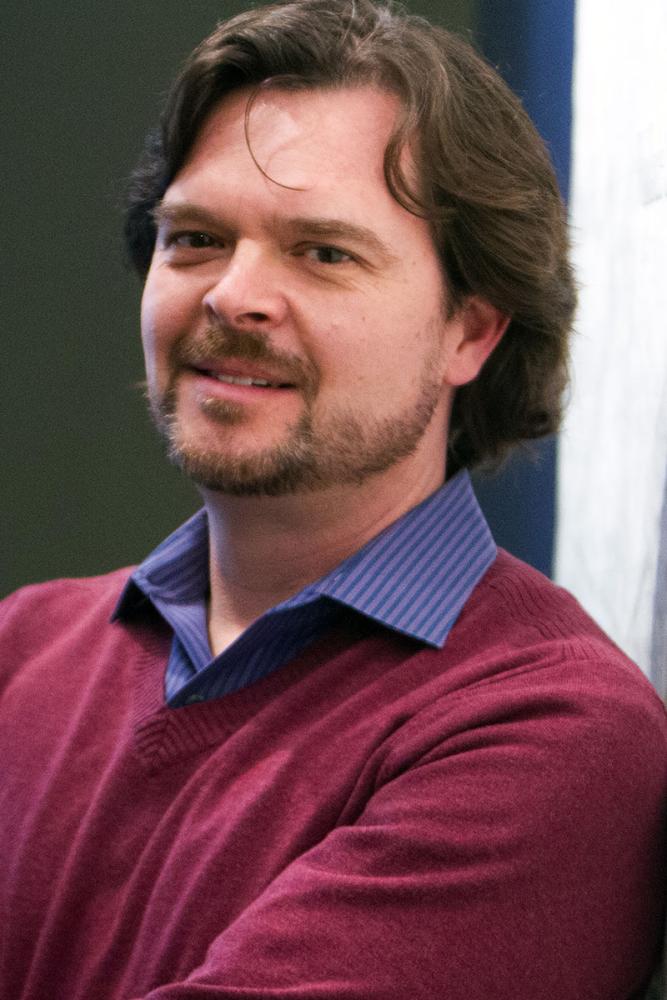 Leonard Nevarez with short brown hair and beard wearing a maroon sweater over a blue collared shirt, smiling with arms crossed.