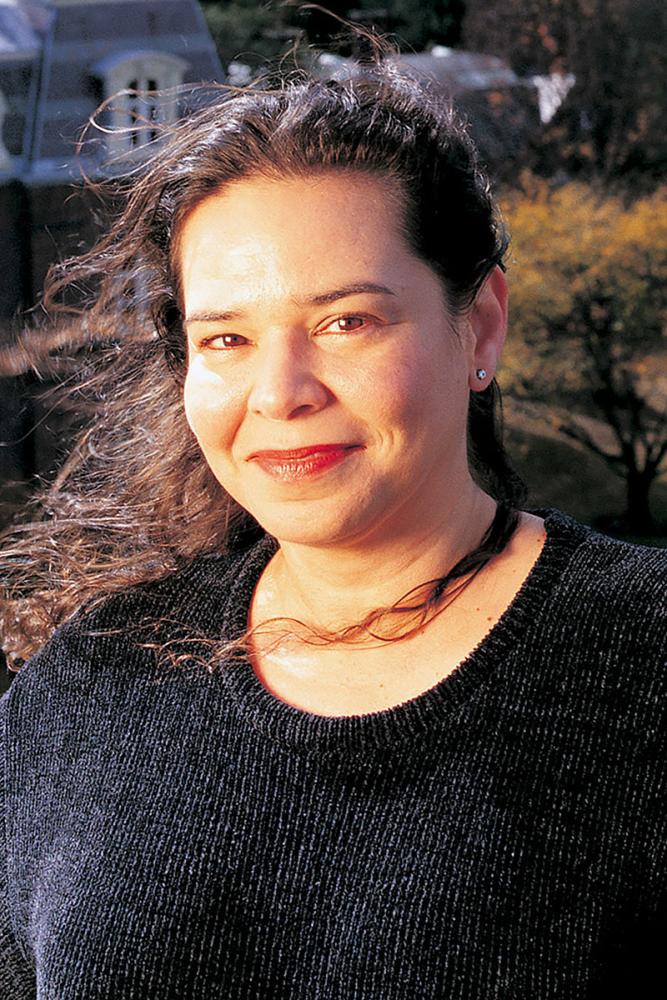 Lizabeth Paravisini-Gebert with long dark hair wearing a dark sweater, smiling outdoors with buildings in the background.