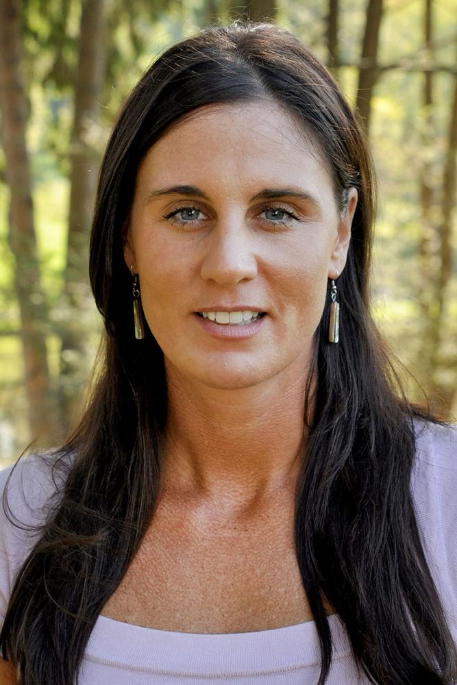 Molly McGlennen has long dark hair and blue eyes, smiling slightly wearing a purple shirt outdoors in a wooded setting.