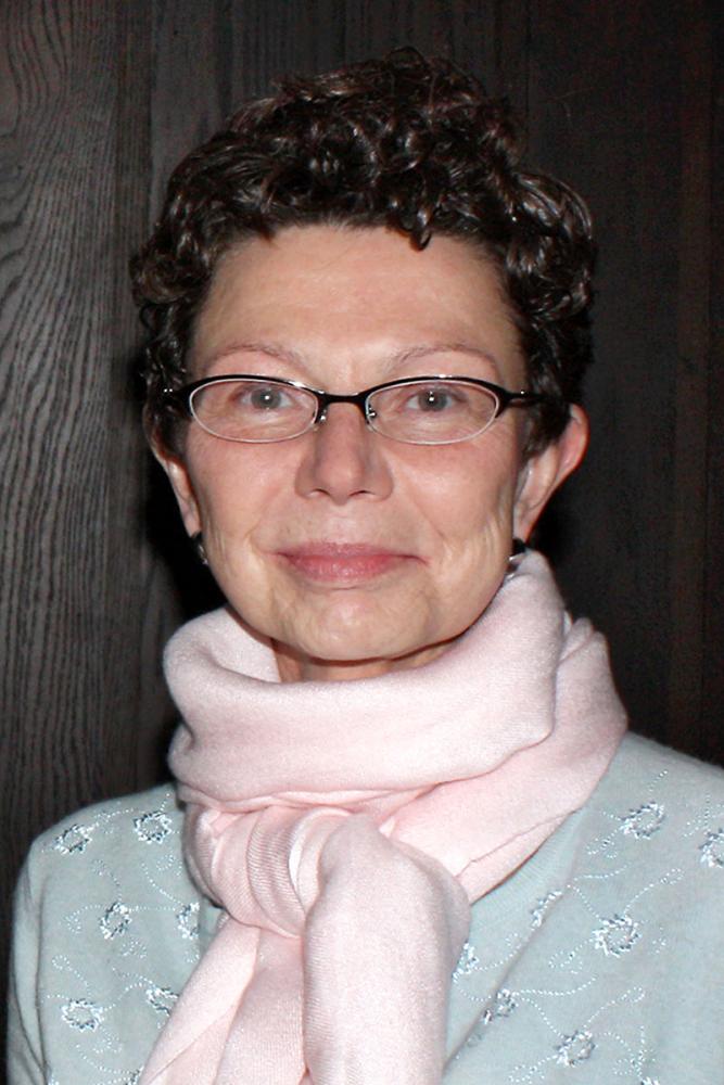 Pinar Batur with short curly hair and glasses wearing a light blue sweater and pale pink scarf, smiling softly indoors.