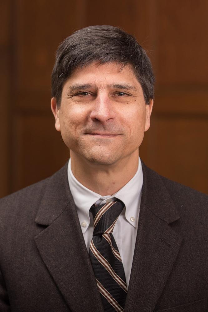 A person wearing a light colored shirt and brown jacket and tie smiles at the camera. They are in a room with wood paneling in the background.