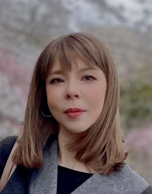 Head and shoulders portrait of a person with long hair and bangs outside with a grey v-neck jacket and black shirt.