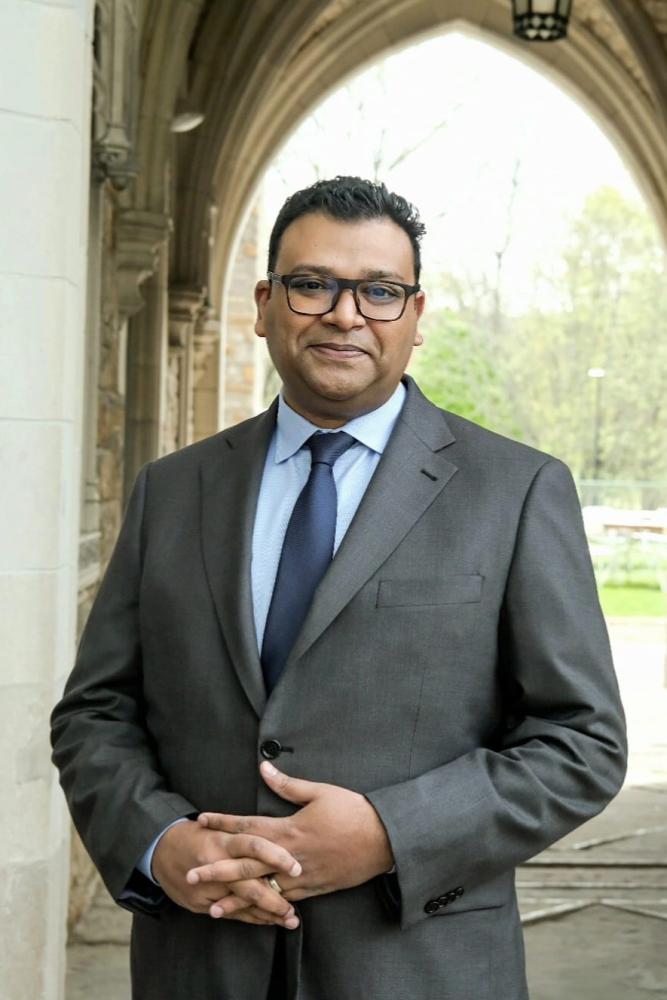 Person in a grey suit with a blue tie standing outside with a archway in the background.
