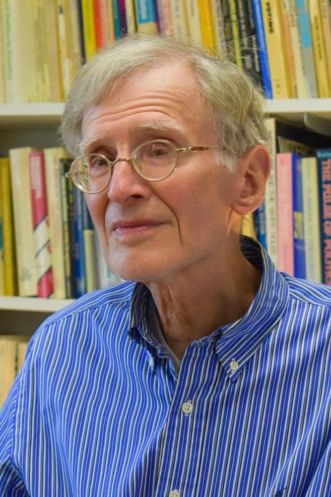 A person wearing glasses and a blue striped shirt sits in front of a bookshelf filled with books.