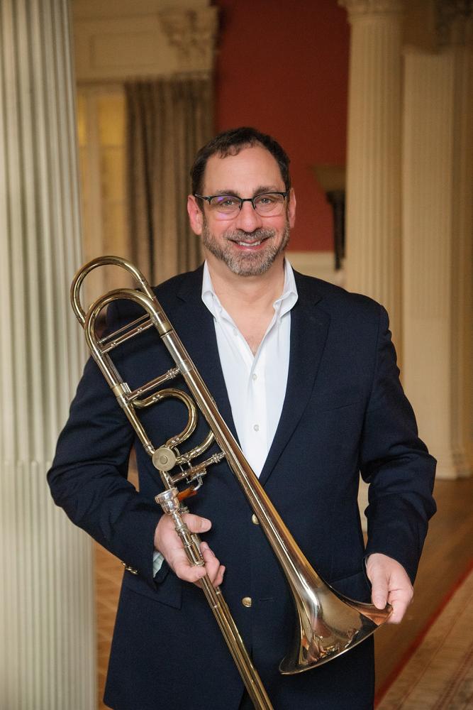 A person in a dark suit jacket and white shirt smiles while holding a brass trombone in an elegant room with tall columns and warm lighting.