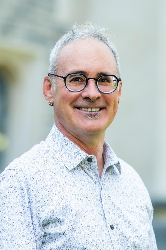 Portrait of a smiling individual in glasses, wearing a light patterned shirt, standing outdoors.