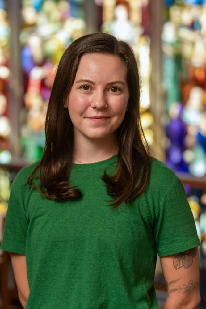 Person in a green shirt standing in the Vassar Library in front of a stained glass window.