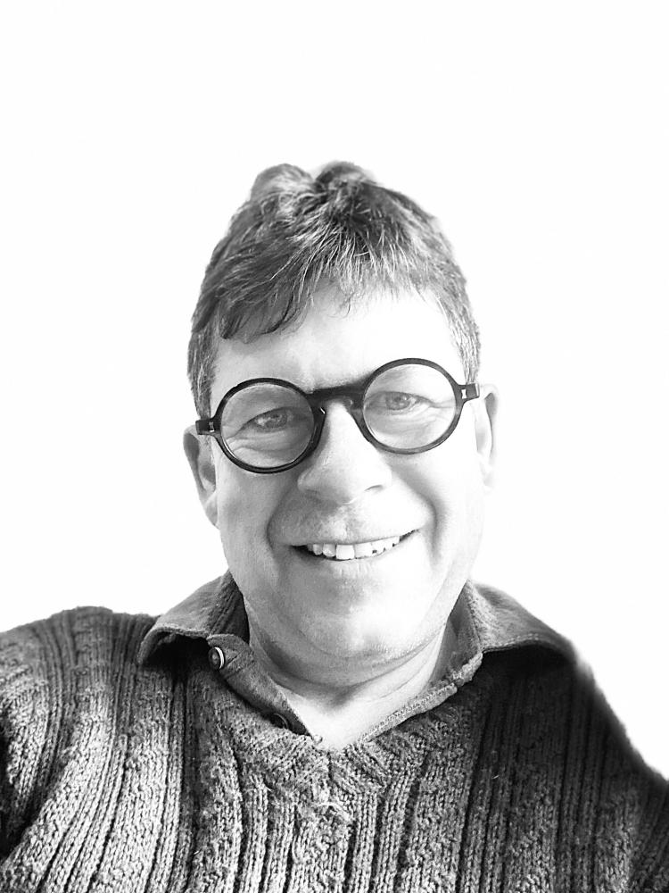 A grayscale portrait of a person wearing round glasses and a textured knit sweater, photographed in black and white against a bright background.