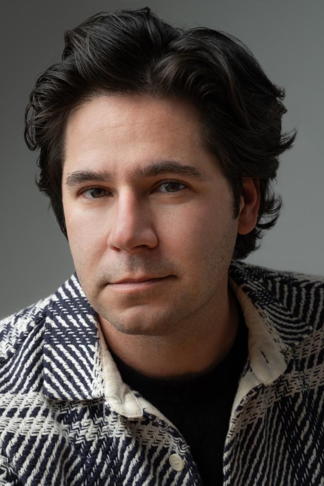 A close-up studio portrait of a person with dark hair, looking directly at the camera with a neutral expression. The person is wearing a dark sweater and a jacket with a black and white chevron pattern. The background is a plain gray.