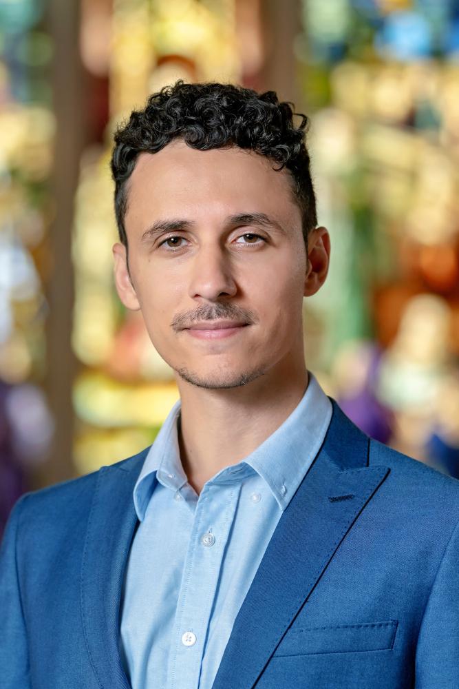 Alan Hankers in a blue suit and light blue shirt stands indoors, looking at the camera with a neutral expression. A colorful stained glass window is blurred in the background.