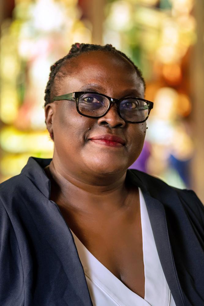 A photo of Patricia Achiro Olwoch, a person with short black hair, glasses, and a black jacket.