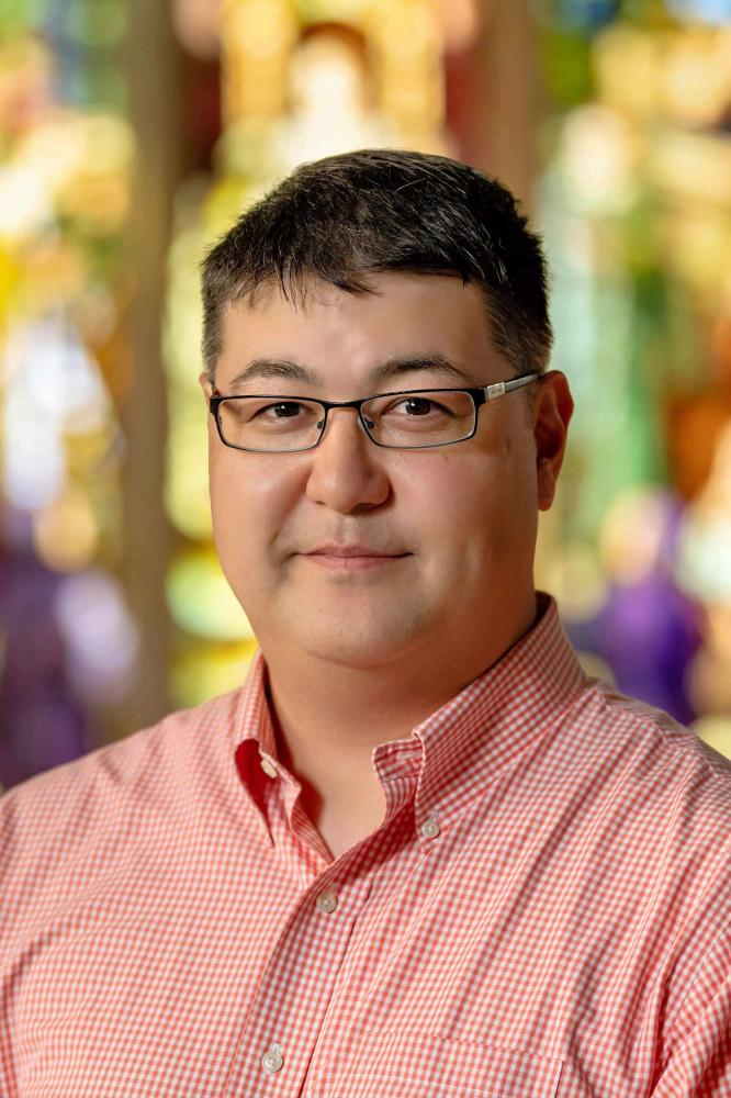 A person with short, dark hair and glasses smiles faintly at the camera. The person is wearing a red and white gingham collared shirt. The background is an abstract blur of warm, bright colors, suggesting a soft focus on stained glass or a vibrant natural setting.