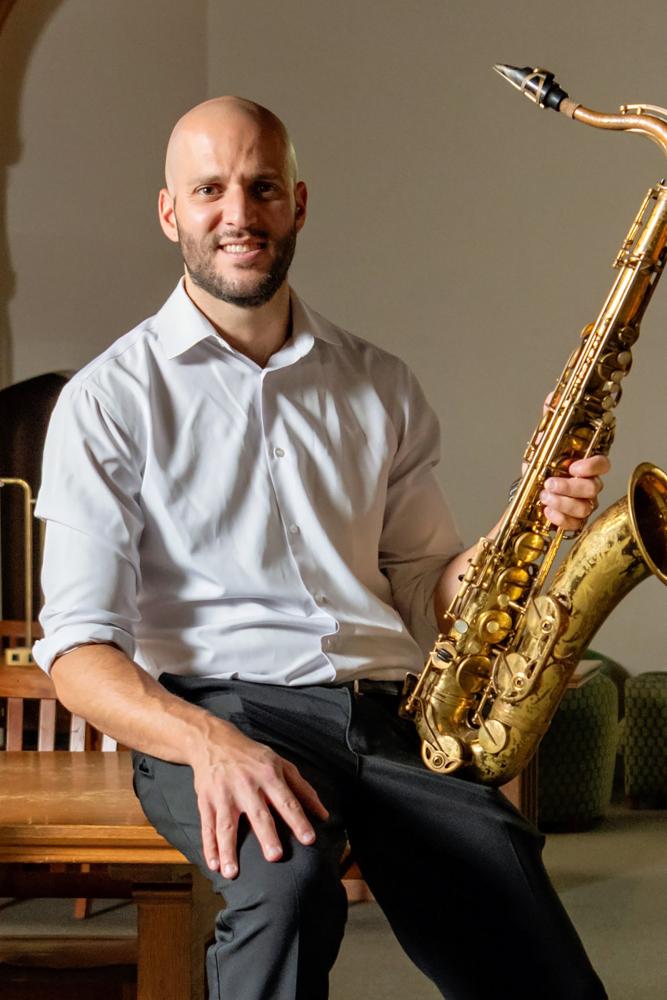 Joseph North in a white dress shirt and dark pants sits indoors holding a saxophone, smiling at the camera.
