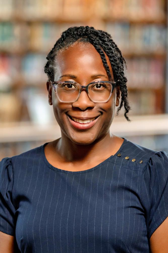 Kimberly Williams Brown wearing glasses and a navy blue pinstriped top smiles while standing indoors, with blurred bookshelves in the background.