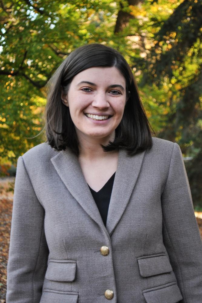 Heather Newman with straight brown hair wearing a gray blazer and black top, smiling outdoors with trees and autumn foliage in the background.