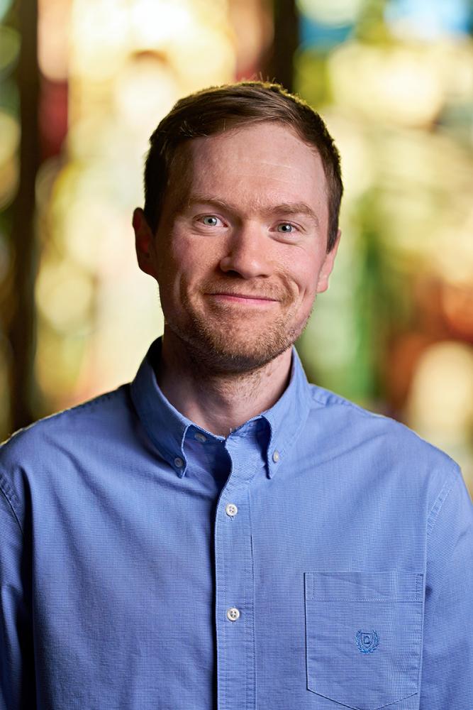 Head-and-shoulders portrait of a person with short light brown hair and light-colored eyes, smiling gently at the camera. They are wearing a blue button-down shirt with a small embroidered logo on the chest pocket. The background is softly blurred and colorful, resembling stained glass or bright abstract panels, creating a warm, vibrant feel.
