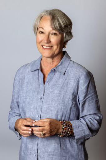 Susanne D. Lyons smiling, and is wearing a light blue linen shirt and a colorful beaded bracelet with with short, styled grey hair and her hands loosely clasped in front of her against a neutral grey background.
