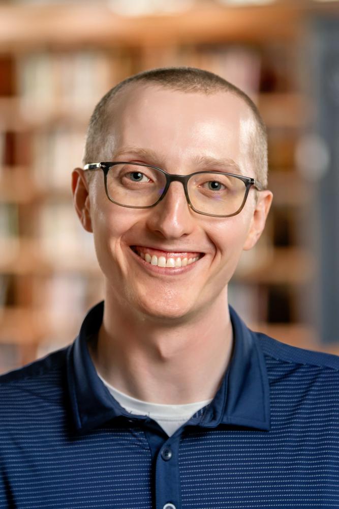 Jacob Erickson smiling with glasses in a blue polo shirt, posing in front of a blurred bookshelf background.