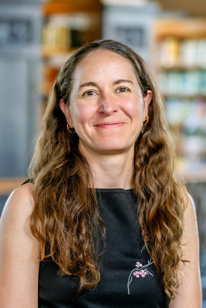 Colette Salyk, with long, wavy brown hair and gold hoop earrings smiling, wearing a black top with subtle pink floral embroidery in front of a soft-focused library background filled with wooden bookshelves.