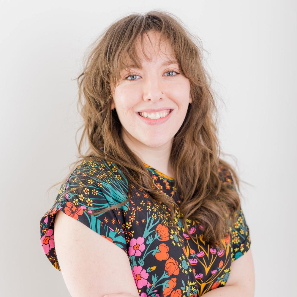 Maya Van Nuys with long wavy brown hair, bangs, and blue eyes, smiling warmly with arms crossed while wearing a black short-sleeved top with a vibrant colorful floral pattern.