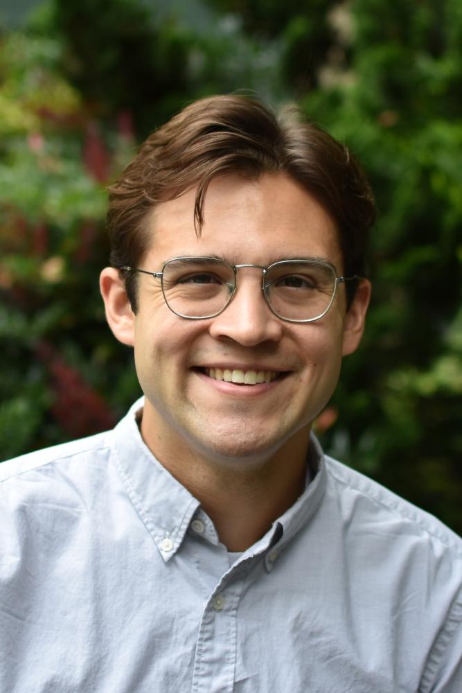 Smiling person wearing glasses and a light-colored collared shirt outdoors.