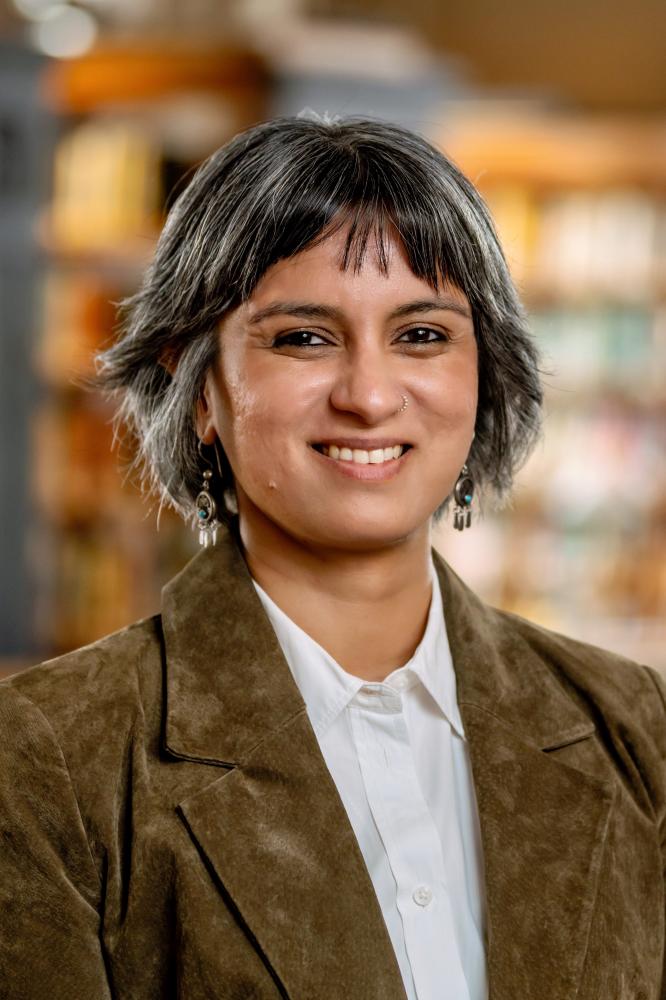 A smiling person with short gray hair wearing a brown blazer and white shirt, with library bookshelves in the background.