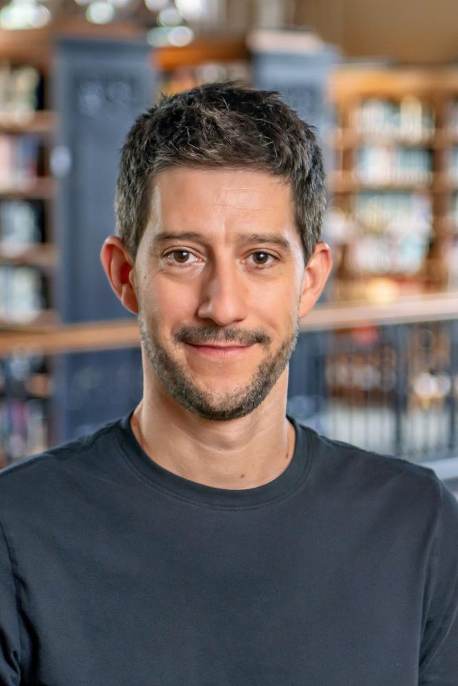 Jesse Delia smiling with short dark hair and light stubble, wearing a plain dark crewneck shirt in a library with wooden bookshelves blurred in the background.