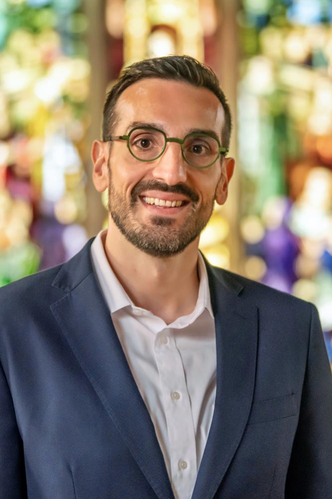 A photo of Mario Sassi, a person with short dark hair, facial hair, glasses, and a dark jacket.