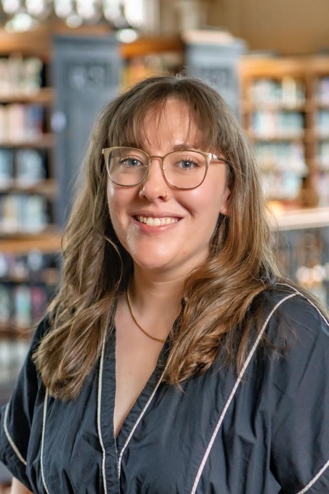 Maya Marie Van Nuys smiling, long wavy brown hair with bangs, and clear-framed glasses, wearing a black top with white piping in front of a softly blurred library background with wooden bookshelves.