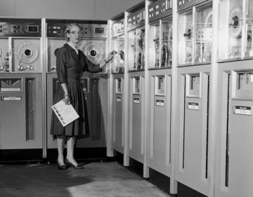 Black and white photo of computer pioneer Grace Murray Hopper, Vassar class of 1928, who developed the computer language COBOL, standing in front of a bank of old tape computers. 