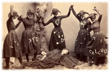 Old black and white photo of five people dancing in black dresses with math symbols and hats with face masks around two people lying down.