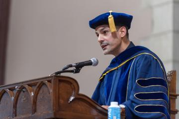 Carlos Alamo-Pastrana speaks in front of a podium. Alamo-Pastrana is wearing a blue robe and blue cap with gold tassel.