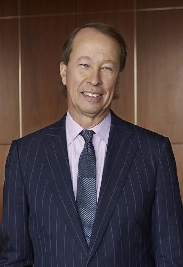 A dark-haired person wearing a suit and tie smiles at the camera.