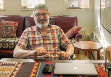 Man with glasses, a beard, and plaid shirt holding a pencil in both hands, sitting in a brightly lit casual office with a sofa and windows in the background. A table with a computer and phone sit in front.