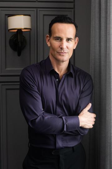 Dr. Paul Frank ’91 wearing navy blue shirt, black pants and standing in front of a gray paneled wall with a small light. 
