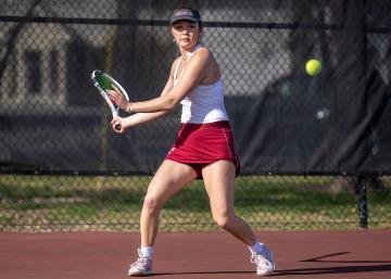Person in Vassar tennis uniform preparing to hit a forehand tennis ball on a red tennis court with a fence in the background.