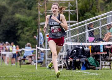 Person running with clear effort in short and t-shirt passing bleachers and spectators next to a running track.