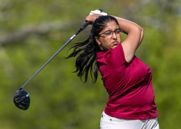 Woman in a Vassar maroon shirt and glasses completing a golf swing with the club and hands above her head.