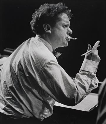 Dylan Thomas, from his right side, wearing a striped collared shirt, holding a pencil in his right hand and a cigarette in his mouth, in black and white. 